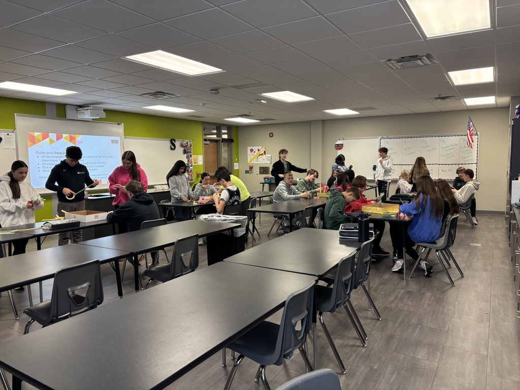 Photo of a classroom full of students. They are gathered around tables working together to build roller coasters.