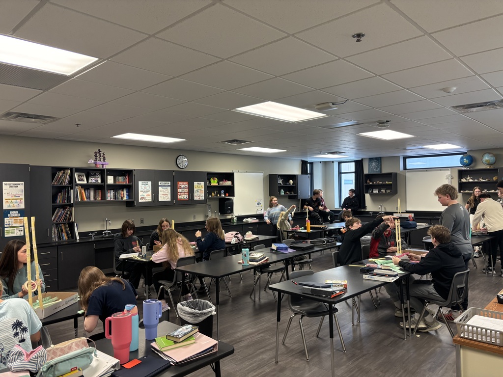 Photo of a classroom full of students. They are gathered around tables working together to build roller coasters.