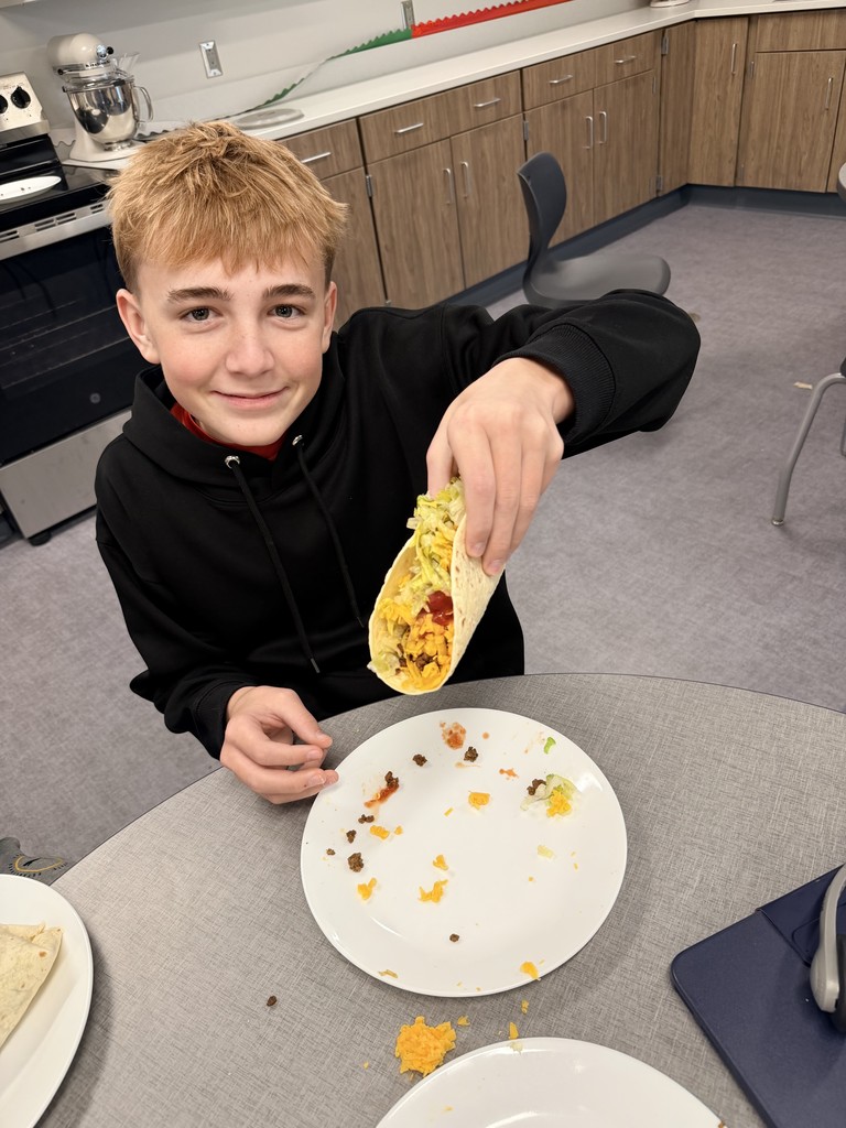 A male student sits at a table and is holding a taco up to the camera.