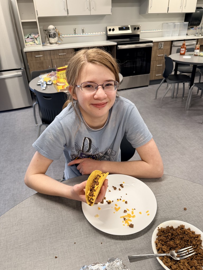 A female student sits at a table with a hard shell taco in her hand. She is smiling at the camera.