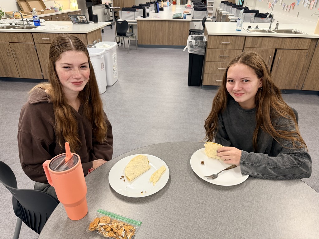 Two female students sit at a table with tacos on a plate between them.