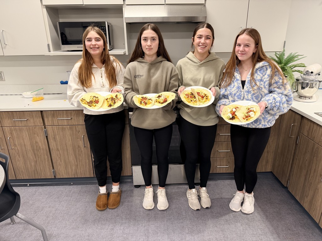 A group of 4 female students stand in a kitchen and hold plates of tacos to the camera.