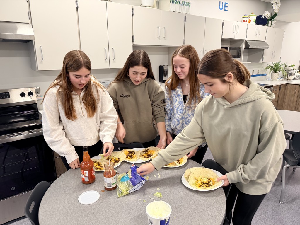 Four female students are at a circular table holding plates and adding lettuce to their tacos.