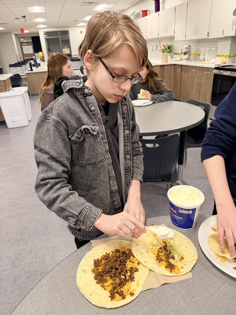 A male student stands at a table and adds sour cream to a soft shell taco.