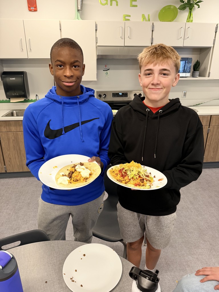 Two male students stand in a kitchen setting and hold plates of tacos.