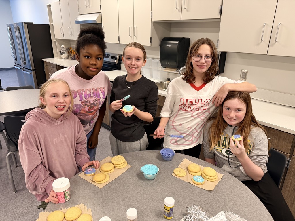 Five female students sit at a round table. All of them are smiling at the camera. They have cookies in front of them and bowls of frosting and are starting to decorate them.