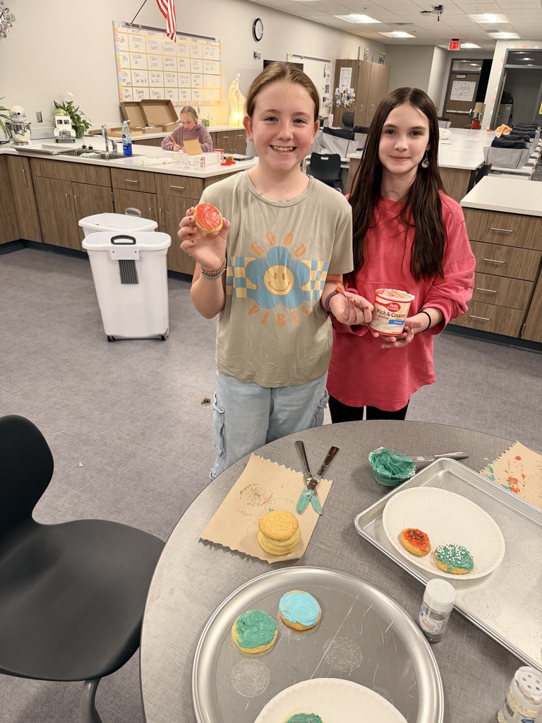 Two female students stand by a round table with cookies and frosting on it. They are holding a cookie and a jar of frosting and are smiling at the camera.