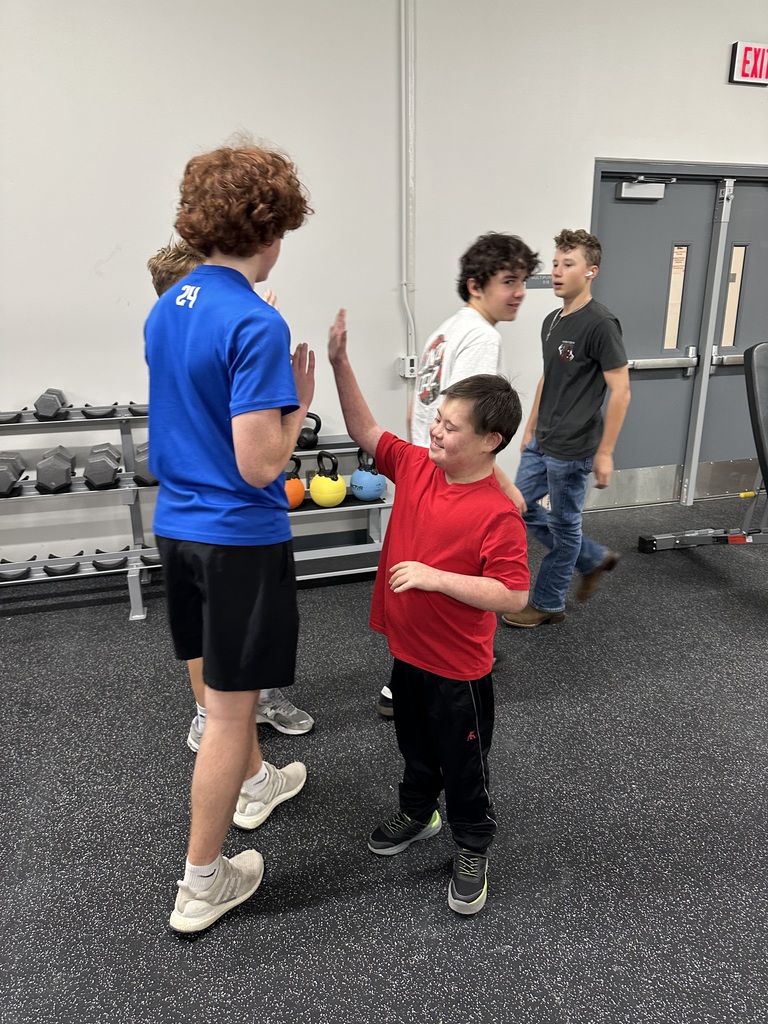 Five male students are in a weight room. One boy with a blue shirt is giving another boy (in a red shirt) a high five.