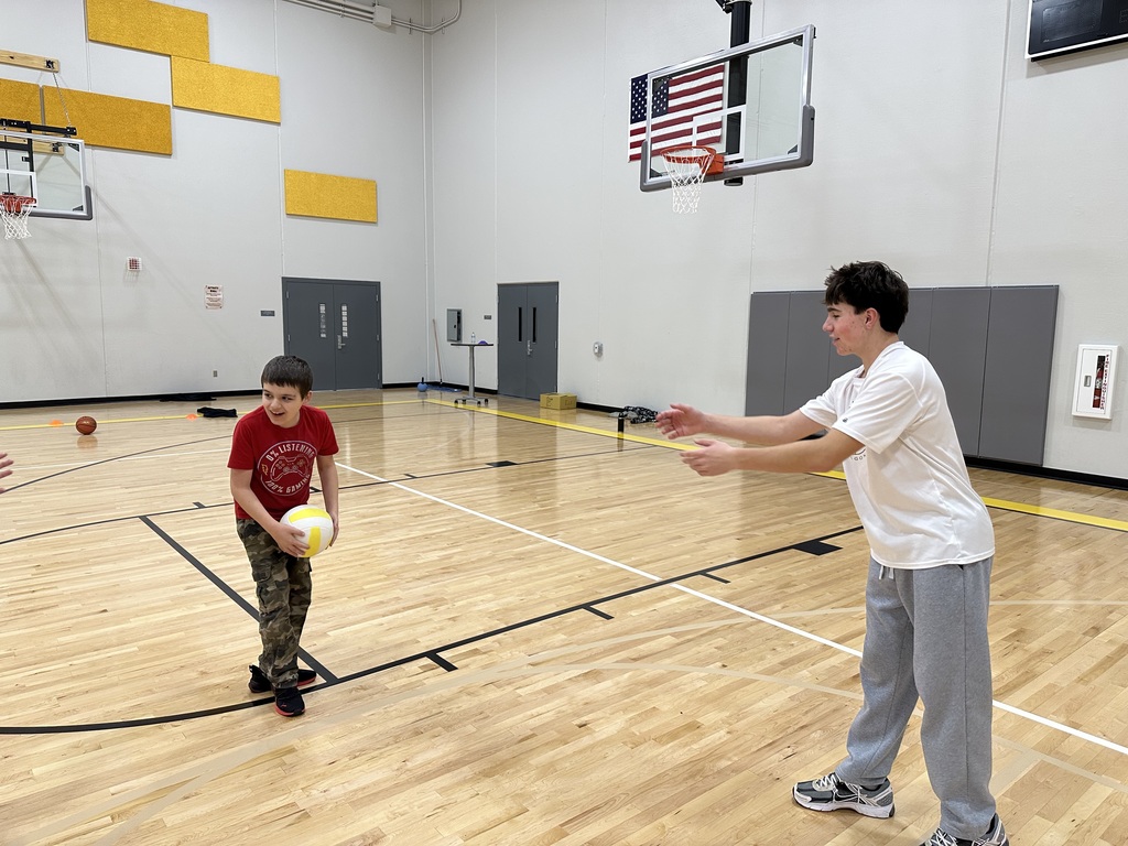 Two male students are in a gymnasium. They are tossing the ball back and forth.