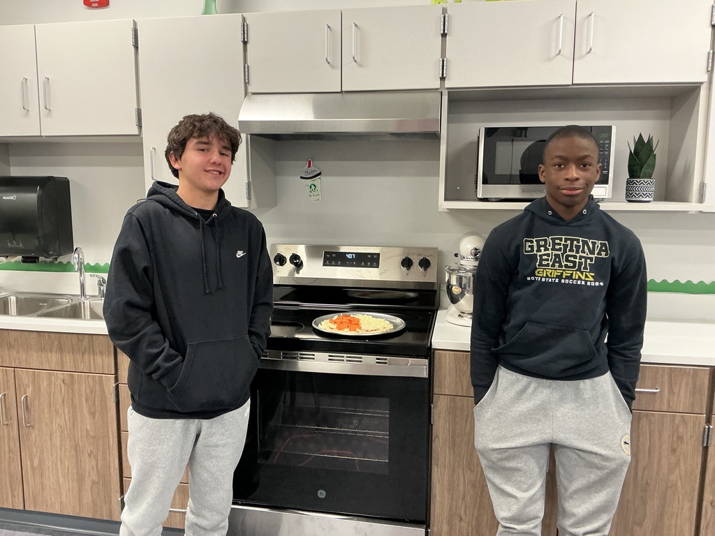 Two male students stand in front of an oven that has an unbaked pizza on it. They are smiling at the camera.