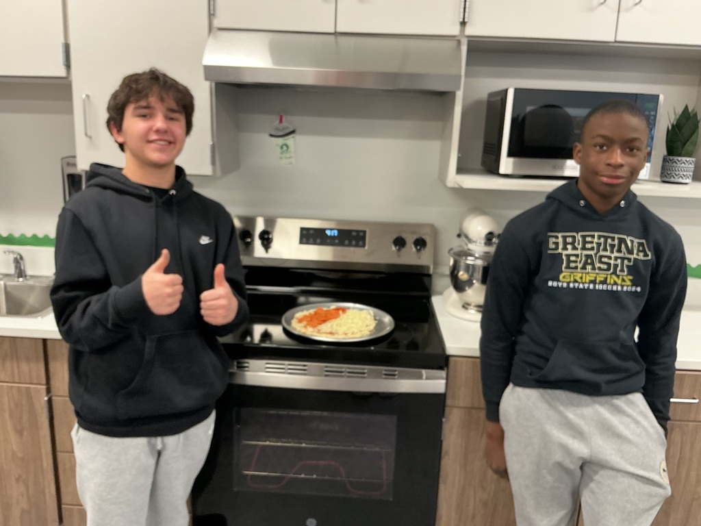 Two male students stand in front of an oven that has an unbaked pizza on it. They are smiling at the camera and the one on the left has his thumbs up.