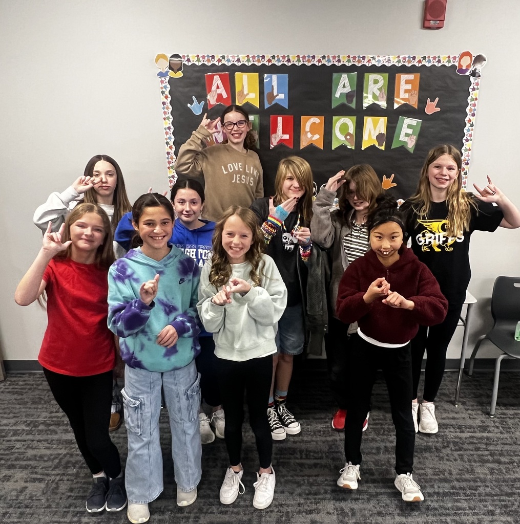 A group of 10 female student stand in front of a black bulletin board. It reads "All Are Welcome" in sign language. They are signing.