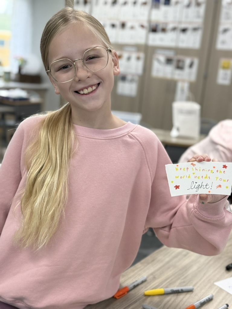 a student holding their decorated kindness coffee sleeve that says "keep shining, the world needs your light!"