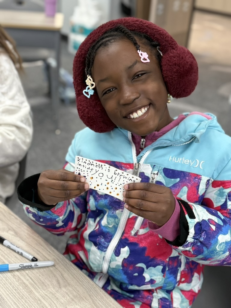 a student in the classroom sitting at table with their decorating coffee sleeve "kindness is brewing inside you!"