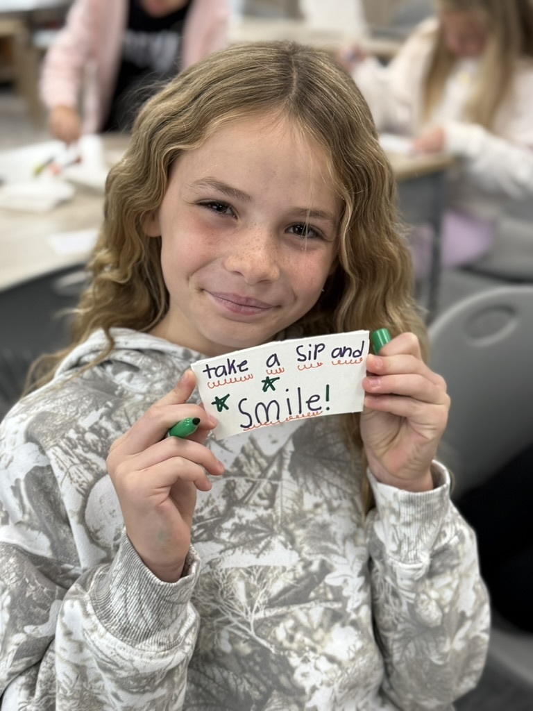 a student smiling at the camera and holding up decorated coffee sleeve which says "take a sip and smile"