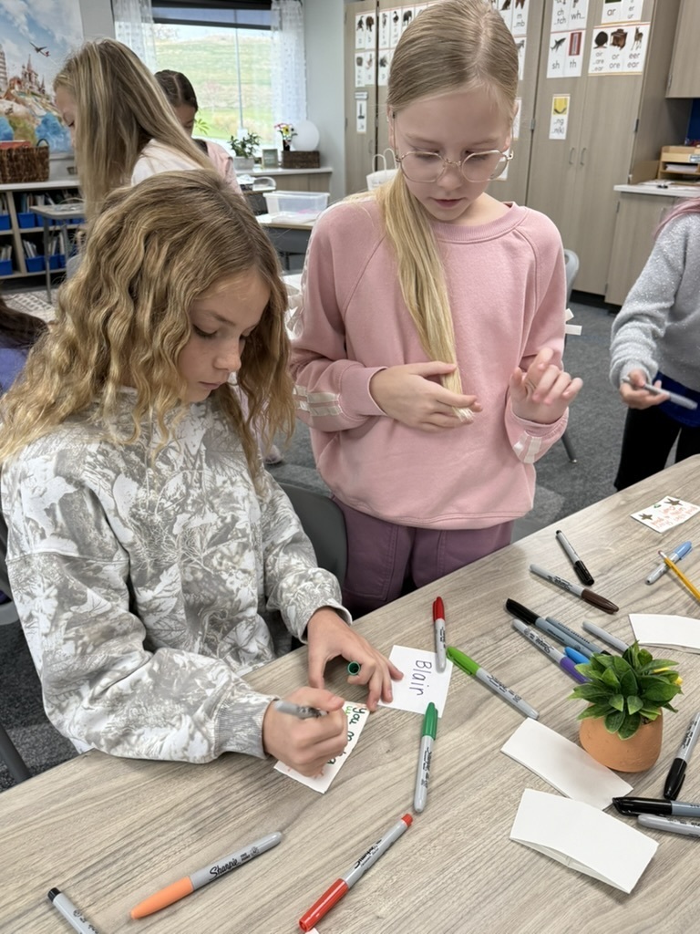 Two students at a desk writing and decorating coffee sleeves for kindness