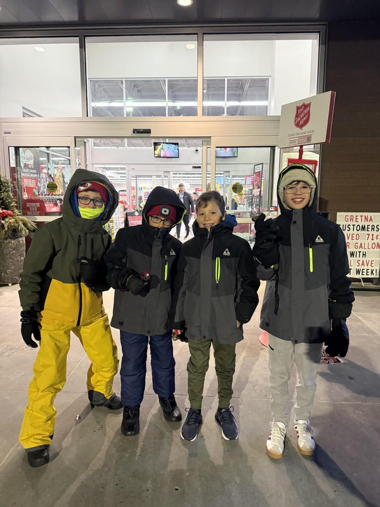 Four young boys in winter coats, gloves, and hats stand smiling in front of an automatic glass doorway of a store with a Salvation Army sign above and to the right of the entrance. The boy on the far left wears a green and yellow jacket, while the other three wear gray and black jackets.