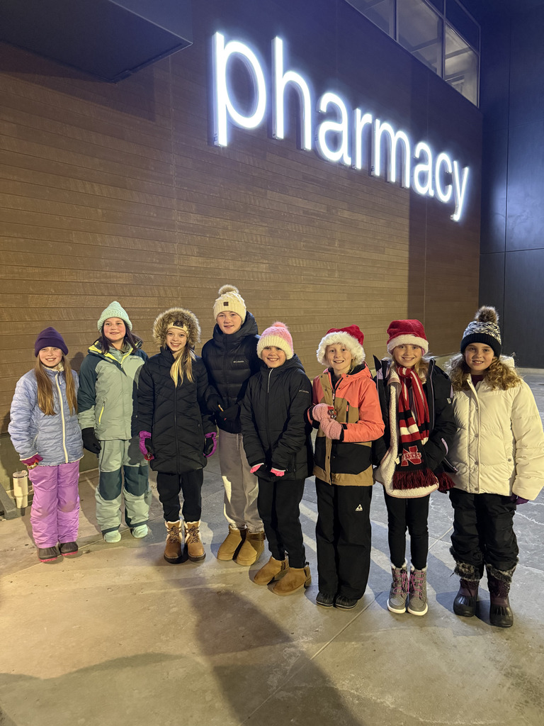 A group of seven children, mostly girls, wearing winter coats and hats stand outside at night beneath a large, illuminated sign reading "Pharmacy" on a brown wooden-paneled wall. Some children are wearing Santa hats.