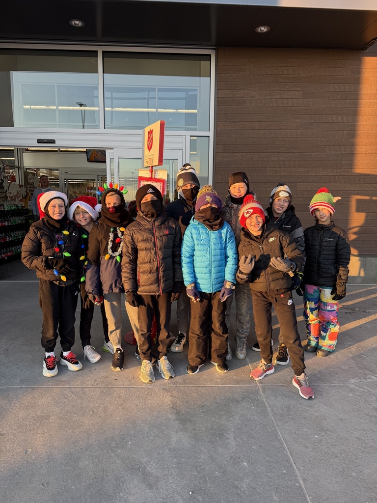 A group of ten children, mostly boys, in winter coats and hats stand together outside during sunset. Several children are wearing string lights or other holiday accessories.