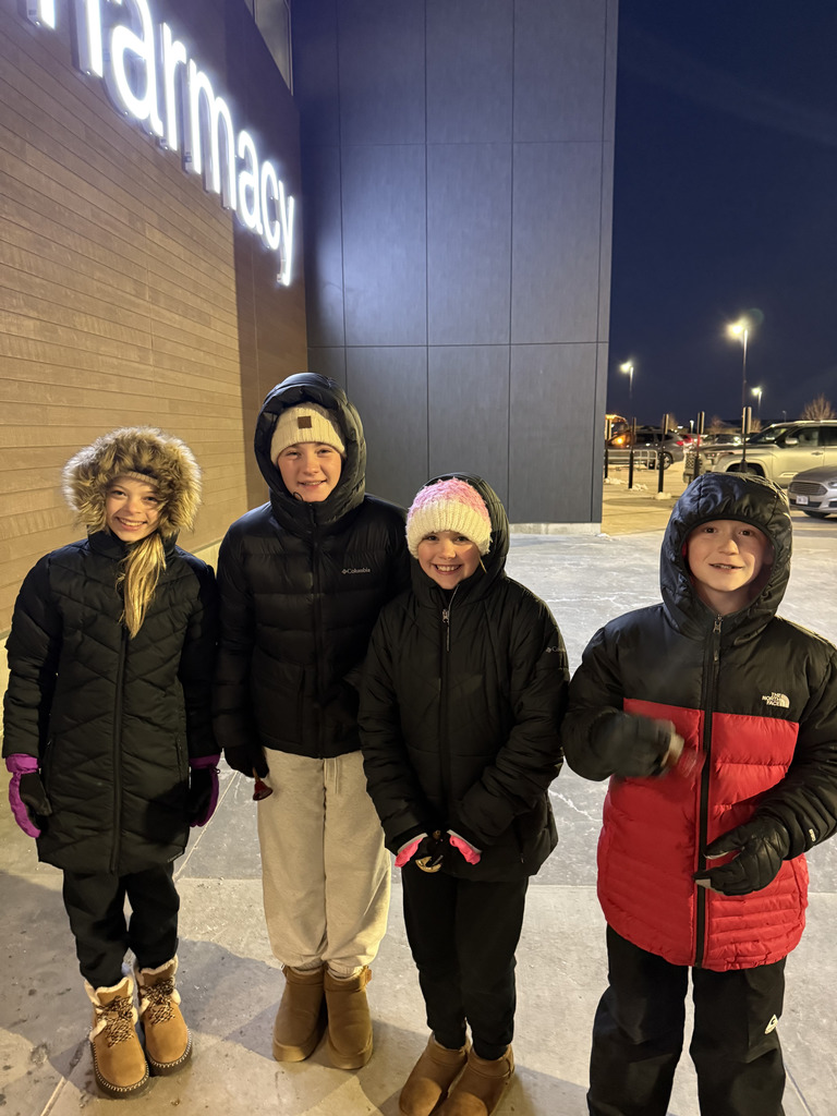 A group of four children—three girls and one boy—stand outside at night in winter coats and hats beneath a large, partially visible, illuminated sign that reads "Pharmacy." The boy on the far right wears a red and black jacket.