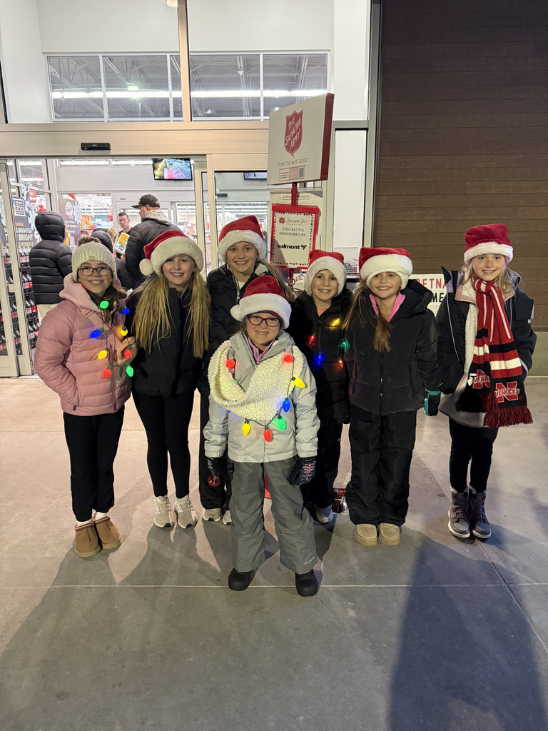 A group of six young girls stands smiling in front of a store entrance. They are all wearing winter jackets and Santa hats. The girls on the left and center are wrapped in colorful holiday string lights, and a Salvation Army donation kettle and sign are visible behind the group.