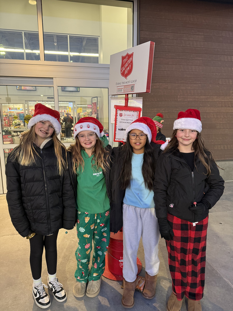 Four girls wearing Santa hats and winter clothing stand smiling in front of a store entrance. A sign for The Salvation Army and a red donation kettle are visible between the girls in the center. The girl second from the right wears a green shirt and Christmas-themed pajama pants, while the girl on the far right wears red and black plaid pajama pants.