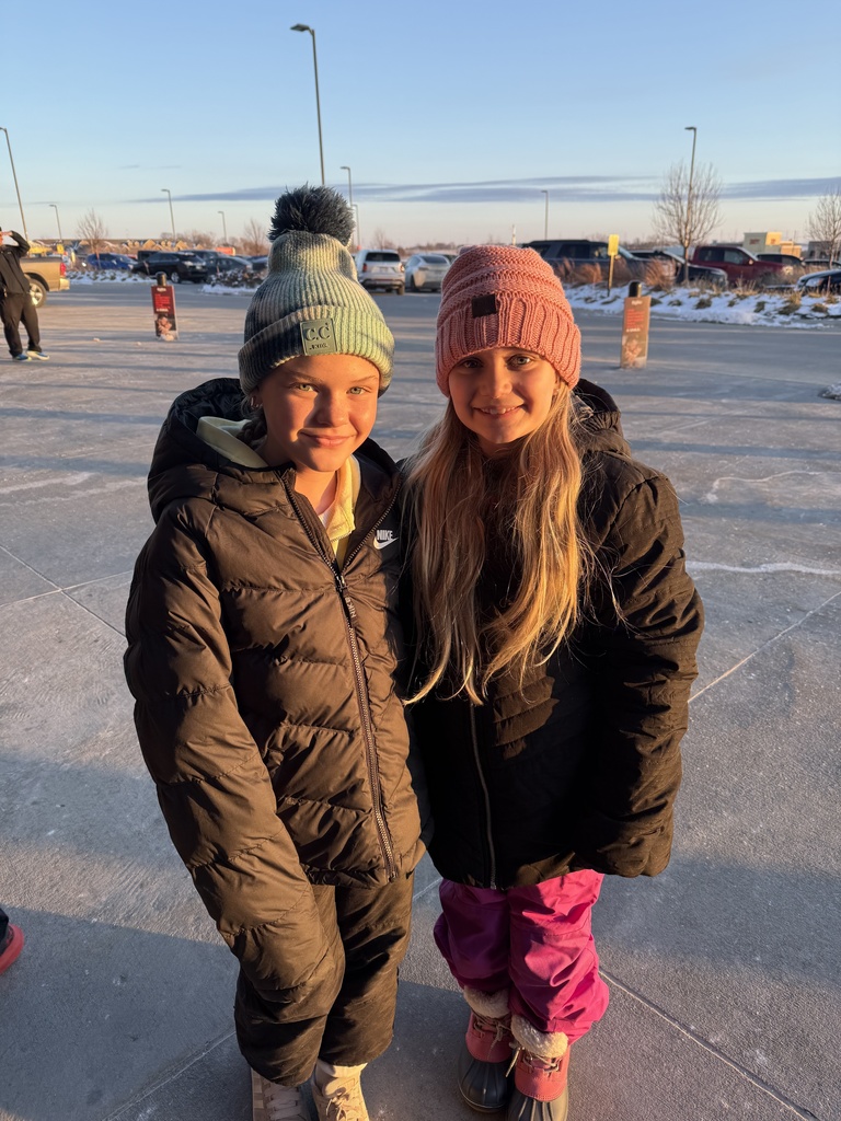 Two young girls in dark winter puffer coats and pom-pom knit hats stand close together and smile at the camera outdoors at sunset. The girl on the right is wearing bright pink snow pants.