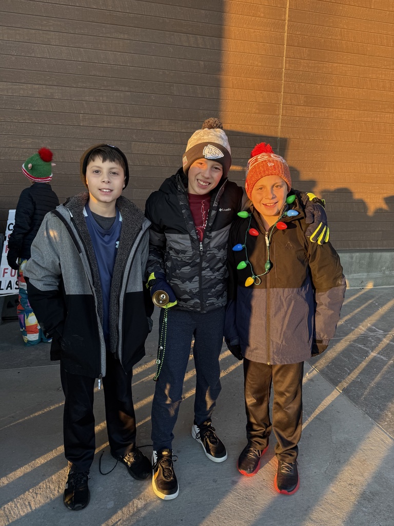 Three boys in winter hats and coats stand side-by-side outdoors at sunset. The boy in the middle holds a bell, and the boy on the right is wrapped in colorful oversized string lights.