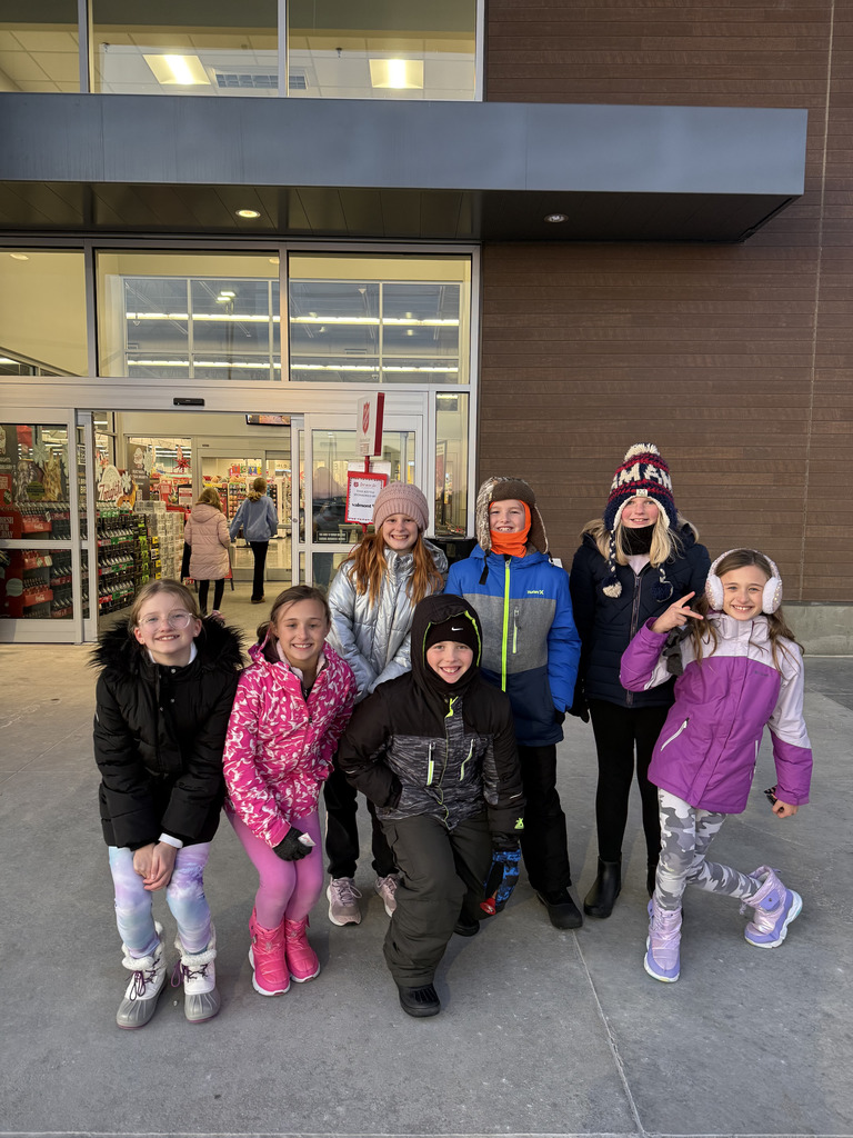 A group of seven children wearing winter coats, hats, and boots stand smiling in front of an automatic glass doorway of a store during the day. A Salvation Army sign is visible in the background near the doors. The child in the center front is kneeling.