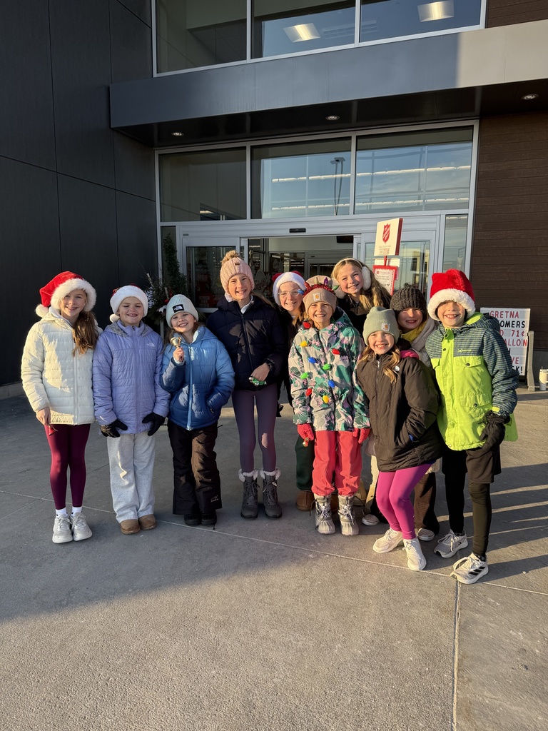 A large group of approximately ten children, mostly girls, wearing winter coats, hats (including several Santa hats), and leggings, stand smiling in front of an automatic glass doorway of a store during the day.