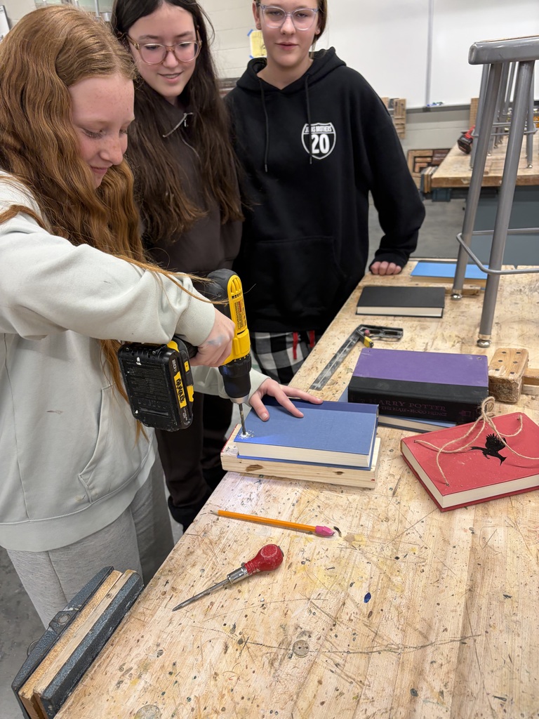 Photo of a female student drilling a hole in her book while two others watch her.