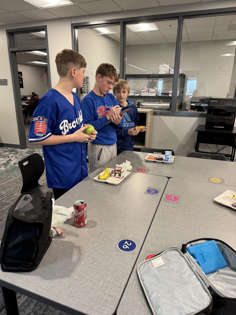 Three boys are standing by a table. The one in the middle has a marker board. They have lunch trays and food in front of them.