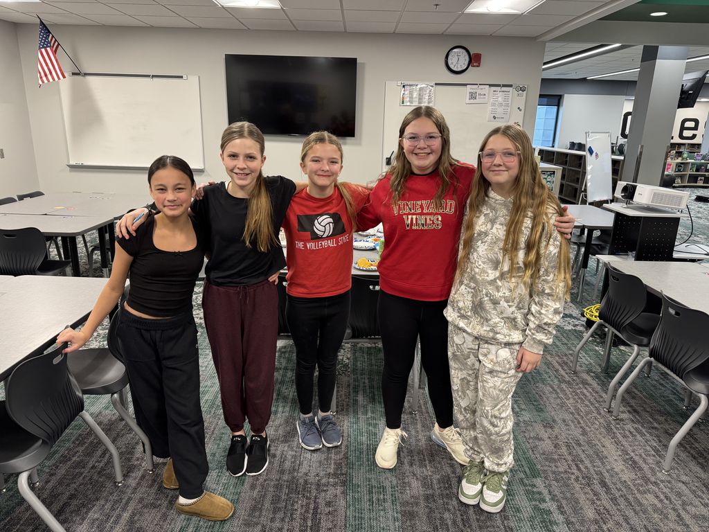 Five girls stand with each other smiling at the camera. They are in a classroom.