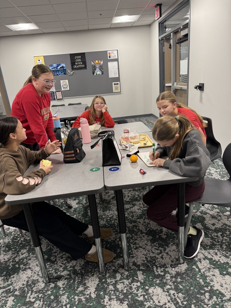 Five girls sit around a table with their lunch trays. They are writing on a small marker board.