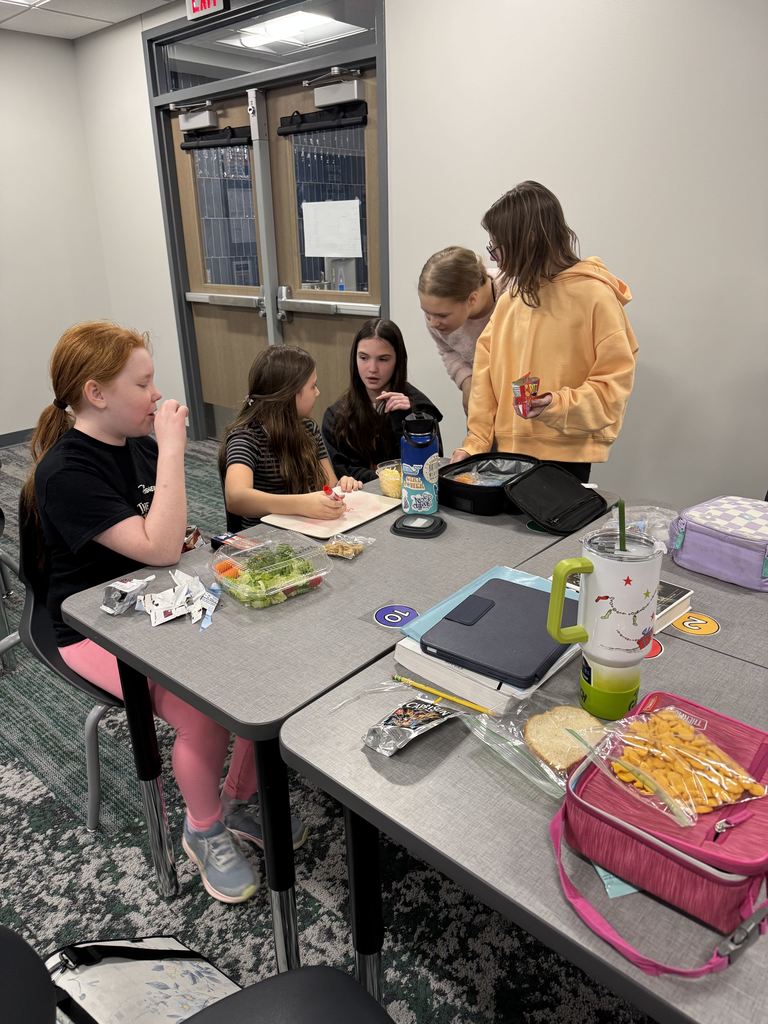 Five girls huddle at the back of a table. They are eating their lunch and discussing an answer. One is writing on a marker board.