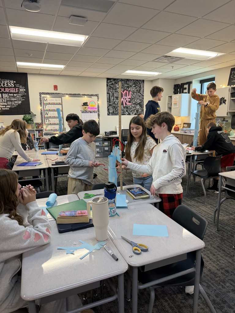 Students in a classroom are gathered around desks. They have paper and measuring sticks out to try to build a tower.
