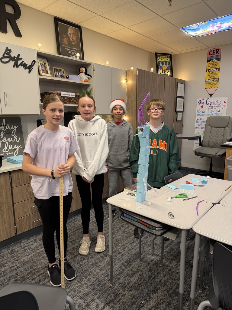 A group of 4 students are smiling at the camera. They are standing at their desks and have a blue paper tower in the middle.
