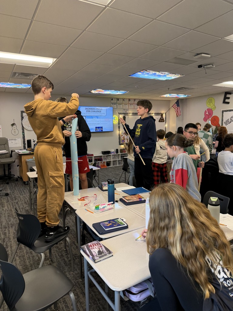 A student stands on a chair next to a group of desks and other students. They are measuring their paper tower.