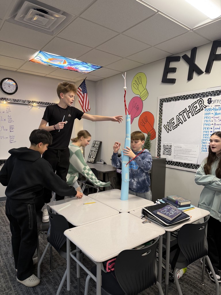 A student stands on a chair next to a group of desks and other students. They are measuring their paper tower.
