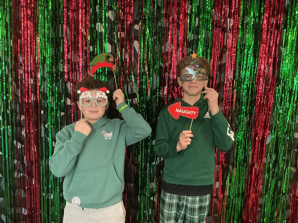 Photo of two male students standing in front of a green and red backdrop holding holiday-themed glasses and hats to their heads and faces. One is also holding an arrow that says naughty and is pointing to his friend.