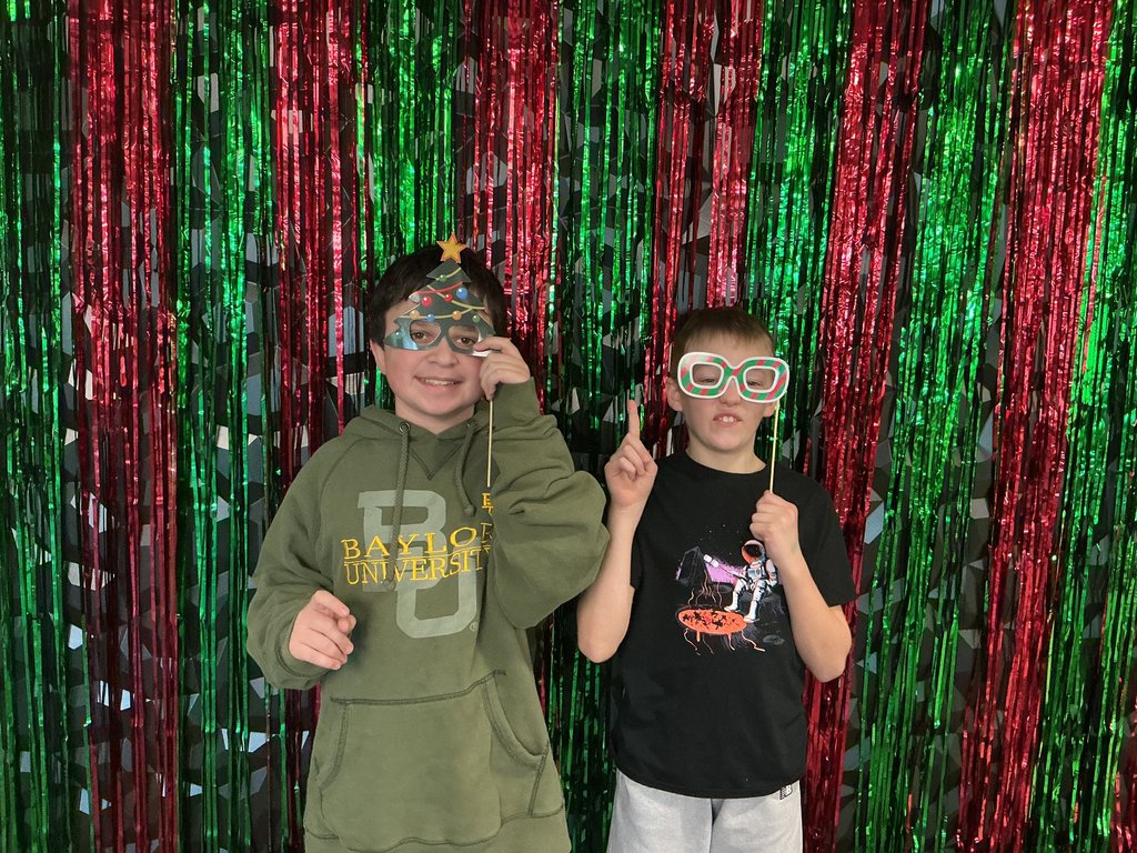 Photo of two male students standing in front of a green and red backdrop holding holiday-themed glasses to their faces.