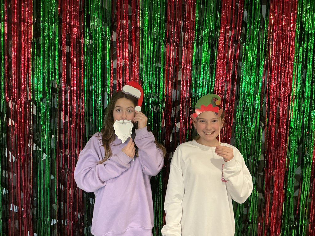 Photo of two female students standing in front of a green and red backdrop holding holiday-themed hats on their heads. One is also holding a beard up to her face.