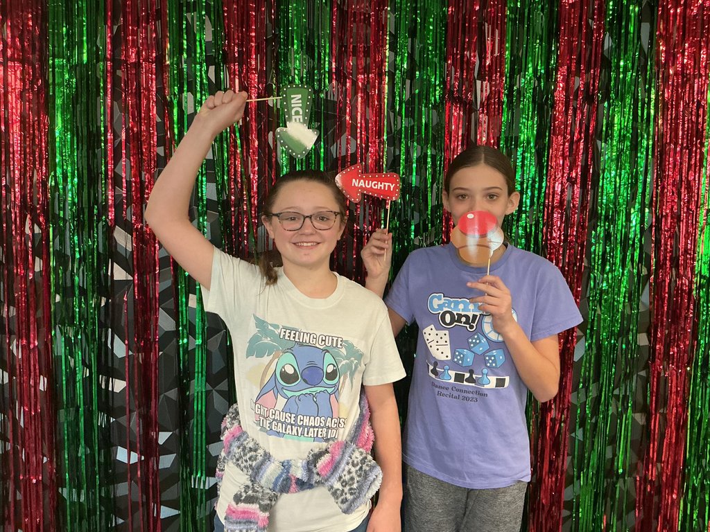 Photo of two female students standing in front of a green and red backdrop holding holiday-themed reindeer noses to their face. One is holding a green arrow that points to herself an says nice. The other is holding a red arrow that points to her friend and says naughty.