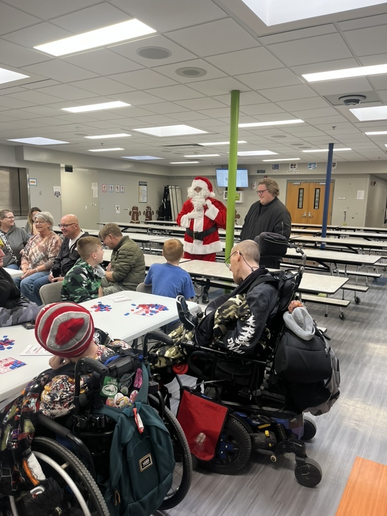 Group of students and adults in a cafeteria sitting at tables with Santa standing in the center of the room.