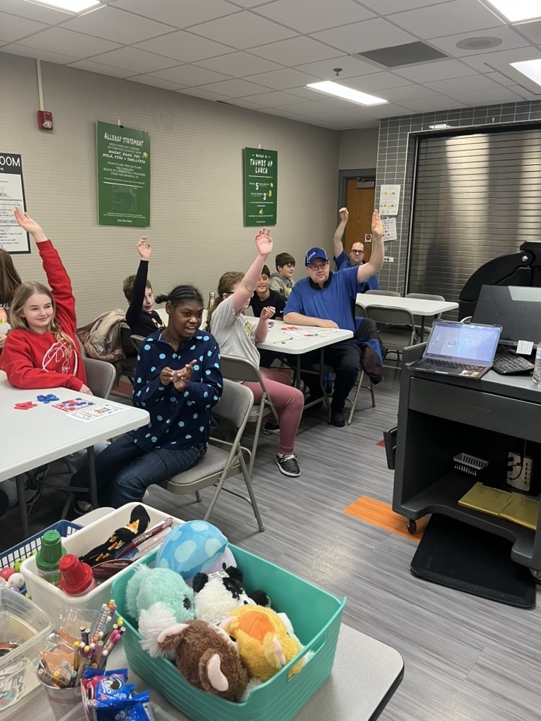 Group of students and adults in a cafeteria sitting at tables playing bingo with their hands raised.