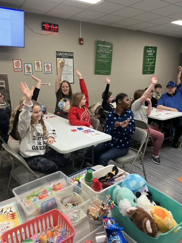 Group of students in a cafeteria sitting at tables playing bingo with their hands raised.