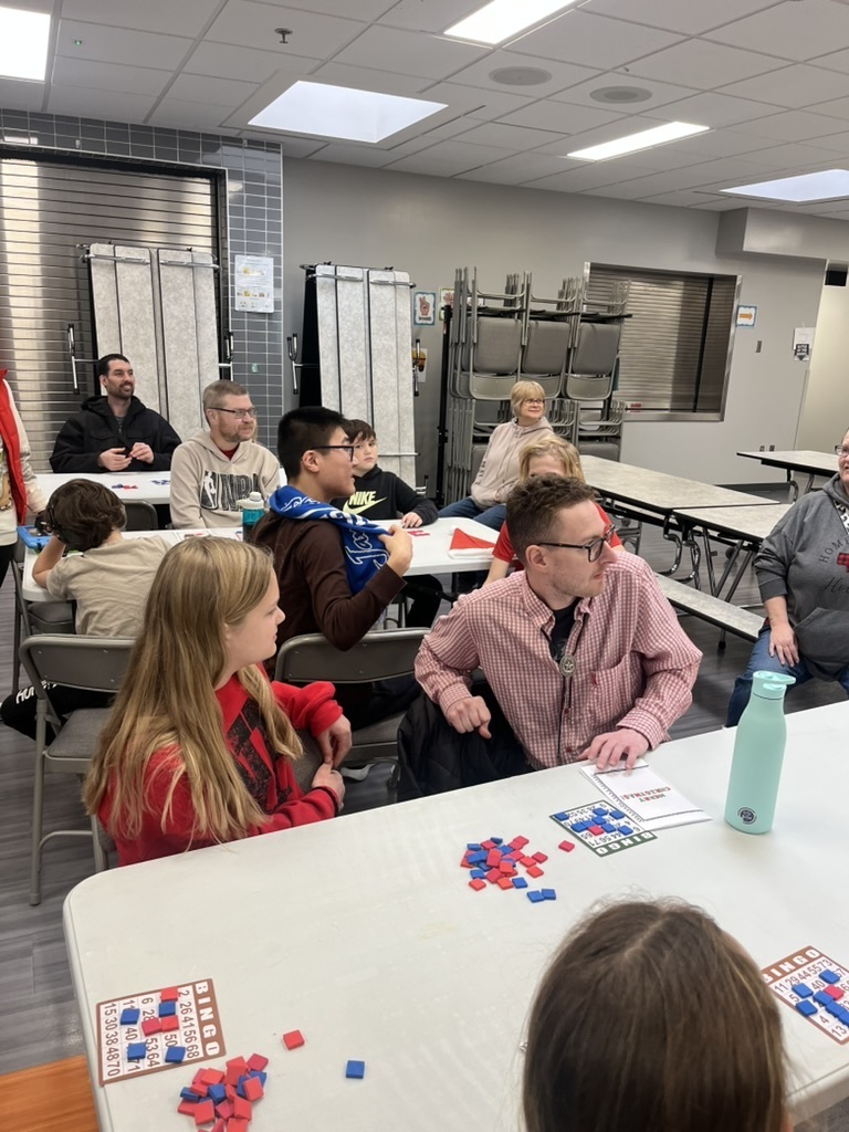Group of students and adults in a cafeteria sitting at tables playing bingo.