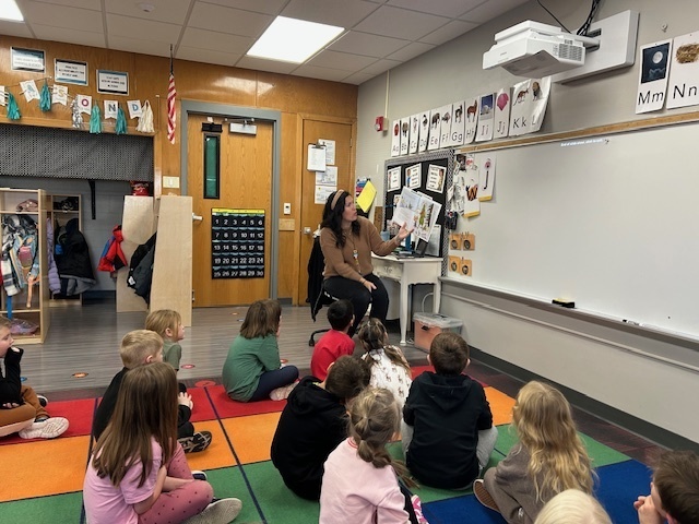 Adult sitting in a chair reading a book in the front of a classroom to students who are sitting on a carpet rug.