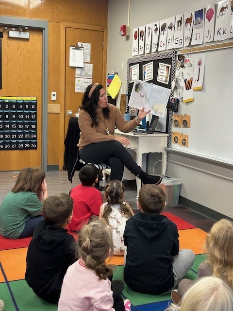 Adult sitting in a chair reading a book in the front of a classroom to students who are sitting on a carpet rug.