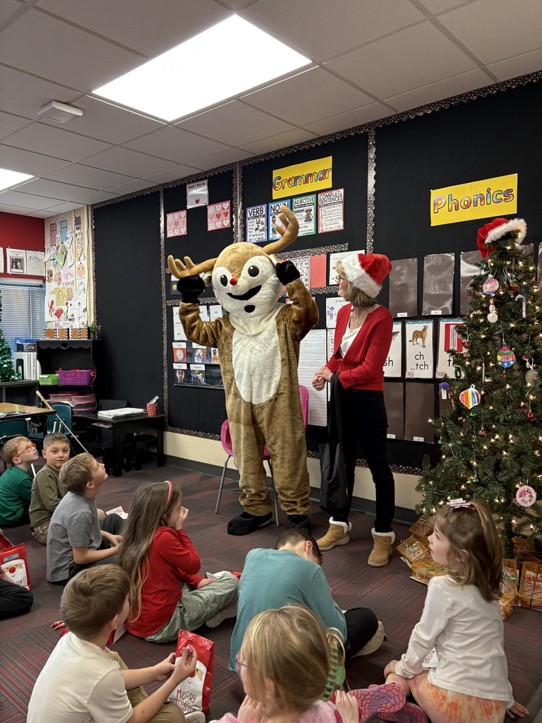 Group of students sitting in a classroom with a person dressed as Rudolph and a woman wearing a red Christmas outfit in the front of the room next to a Christmas tree.